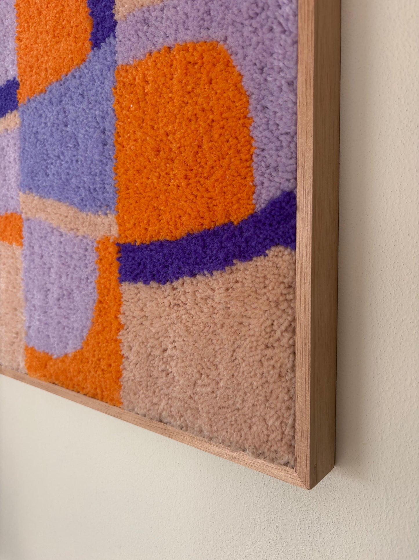 A close-up of a framed abstract wall hanging with textured, looped fibers in orange, lavender, purple, and beige tones, mounted on a light beige wall.